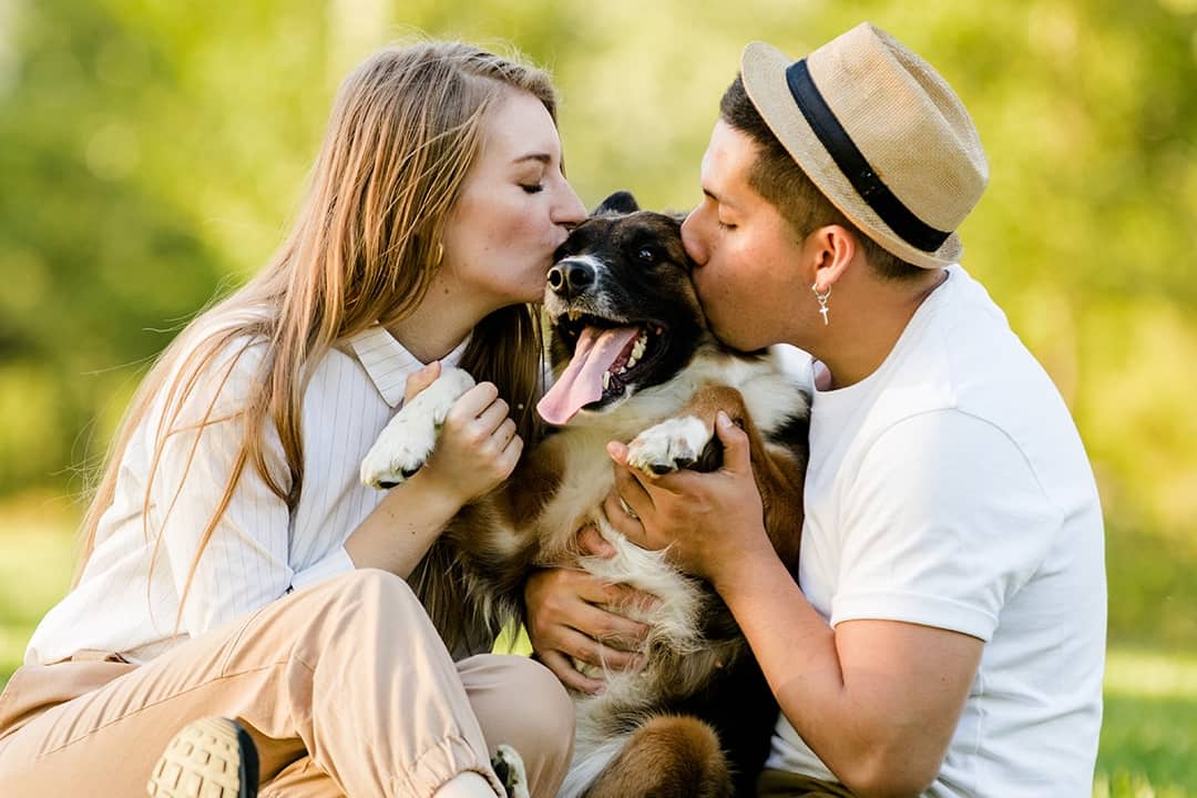 A young couple kissing their pet dog outdoors in a park.